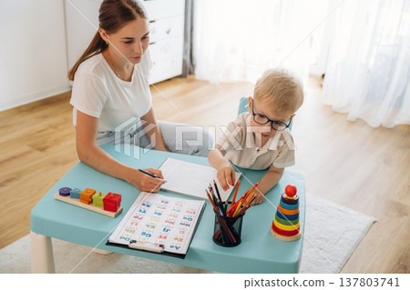 Trying to learn how to draw, holding pencil. Speech therapist is with young boy indoors Trying to learn how to draw, holding pencil. Speech therapist is with young boy indoors 137803741