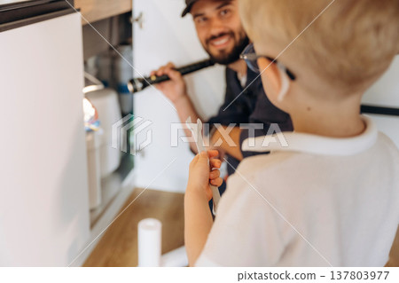 Little boy is helping, holding tool for plumber that is working on the kitchen Little boy is helping, holding tool for plumber that is working on the kitchen 137803977