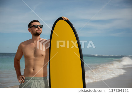 Man stands by the ocean with a surfboard on a sunny day 137804131