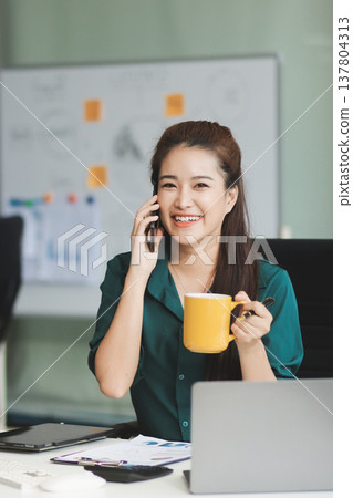 Determined asian businesswoman works late in a modern office, confidently analyzing data on her laptop, embodying success and professionalism with a smile 137804313
