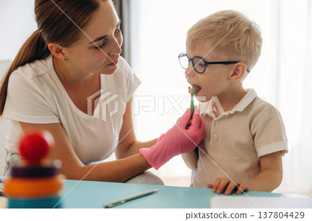 Sitting and doing tongue testing by using special device. Speech therapist is with young boy indoors 137804429