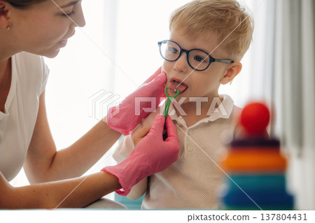 Sitting and doing tongue testing by using special device. Speech therapist is with young boy indoors Sitting and doing tongue testing by using special device. Speech therapist is with young boy indoors 137804431