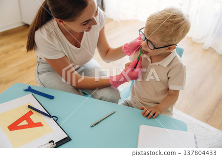 Sitting and doing tongue testing by using special device. Speech therapist is with young boy indoors Sitting and doing tongue testing by using special device. Speech therapist is with young boy indoors 137804433