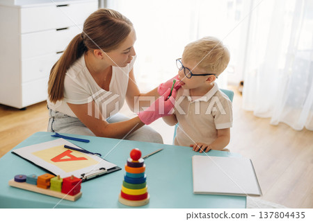 Exercises, using special device on tongue. Speech therapist is with young boy indoors Exercises, using special device on tongue. Speech therapist is with young boy indoors 137804435