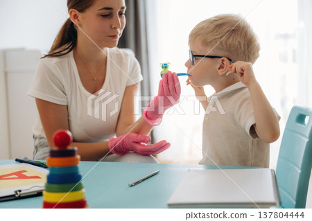 Blows into a tube with little ball. Speech therapist is with young boy indoors 137804444