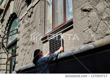 Woman holding photovoltaic solar panel in front of historical building, wearing plaid shirt and sunglasses. Concept of integration of sustainable renewable energy sources into old architecture. Woman holding photovoltaic solar panel in front of historical building, wearing plaid shirt and sunglasses. Concept of integration of sustainable renewable energy sources into old architecture. 137804717