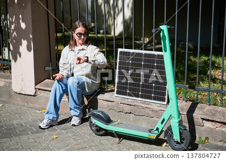 Woman checking her watch while charge electric scooter from portable solar panel, emphasizing integration of sustainable renewable energy. Concept of eco-friendly solutions in urban settings. Woman checking her watch while charge electric scooter from portable solar panel, emphasizing integration of sustainable renewable energy. Concept of eco-friendly solutions in urban settings. 137804727