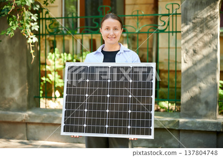 Woman holding solar panel for charging, stands outdoors, smiling at camera, in urban setting. Concept of green energy generation. 137804748