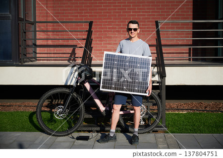 Man holding solar panel for charging, stands in front of a brick building. Cyclist wears sunglasses, light gray shirt, shorts. Next to him pink electric mountain bike with black helmet on handlebars. Man holding solar panel for charging, stands in front of a brick building. Cyclist wears sunglasses, light gray shirt, shorts. Next to him pink electric mountain bike with black helmet on handlebars. 137804751