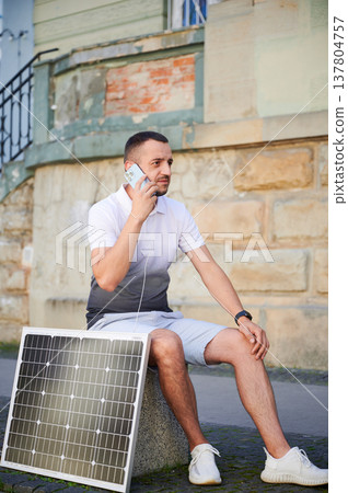 Handsome man using smartphone connected to photovoltaic solar panel. Integration of sustainable renewable energy into everyday life, demonstrating practical use of solar power for charging devices. Handsome man using smartphone connected to photovoltaic solar panel. Integration of sustainable renewable energy into everyday life, demonstrating practical use of solar power for charging devices. 137804757