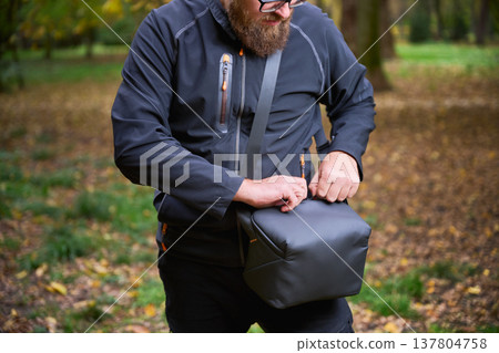 A man dressed in a black jacket is outdoors in a vibrant park. He is busy adjusting his black bag, surrounded by colorful fall leaves and nature. A man dressed in a black jacket is outdoors in a vibrant park. He is busy adjusting his black bag, surrounded by colorful fall leaves and nature. 137804758