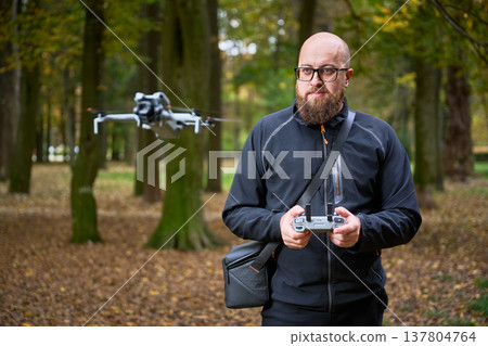 In a peaceful forest during autumn, a man with a beard expertly controls a drone, focused on its flight. Leaves cover the ground, creating a vibrant atmosphere. 137804764