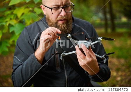 A man with a beard examines and adjusts a drone in a vibrant forest. Leaves turn shades of orange and yellow while sunlight filters through the trees, creating a serene atmosphere. A man with a beard examines and adjusts a drone in a vibrant forest. Leaves turn shades of orange and yellow while sunlight filters through the trees, creating a serene atmosphere. 137804770