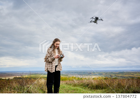 In a serene autumn landscape, a girl stands on a grassy hill, skillfully piloting a drone while surrounded by nature's beauty. In a serene autumn landscape, a girl stands on a grassy hill, skillfully piloting a drone while surrounded by nature's beauty. 137804772