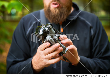 In a lush forest, a bearded man holds a modern device, sharing excitement about technology. Sunlight filters through the trees, enhancing the experience of discovery. 137804774