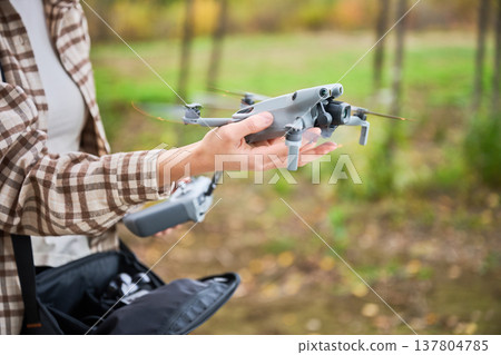 A person holds a modern drone carefully in the woods, surrounded by vibrant autumn leaves and trees, ready for a flight to capture nature's beauty from above. A person holds a modern drone carefully in the woods, surrounded by vibrant autumn leaves and trees, ready for a flight to capture nature's beauty from above. 137804785