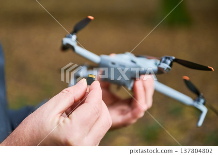 A person is holding a small memory card while standing near a drone in a wooded area. The scene captures the excitement of capturing aerial footage and memories. A person is holding a small memory card while standing near a drone in a wooded area. The scene captures the excitement of capturing aerial footage and memories. 137804802