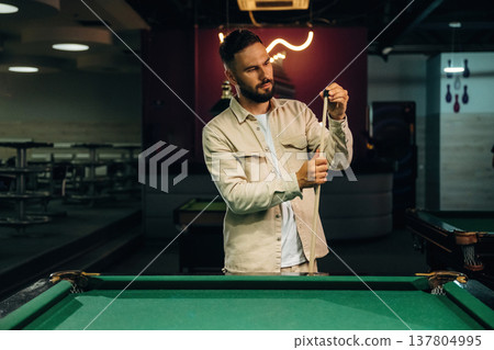 Cleaning up the top of cue stick. Handsome man with a pool table game Cleaning up the top of cue stick. Handsome man with a pool table game 137804995