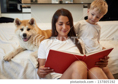 Big red covered book in hands, reading. Mother with son and Shibu Inu dog are at home Big red covered book in hands, reading. Mother with son and Shibu Inu dog are at home 137805192