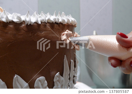 A woman pastry chef with a pastry bag in her hands squeezes cream on a cake in a home kitchen. Professional chef at work on bakery kitchen. culinary, baking and cooking food concept, home cooking A woman pastry chef with a pastry bag in her hands squeezes cream on a cake in a home kitchen. Professional chef at work on bakery kitchen. culinary, baking and cooking food concept, home cooking 137805587