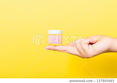 Female hand holding cream bottle of lotion isolated. Girl give jar cosmetic products on yellow background 137805883