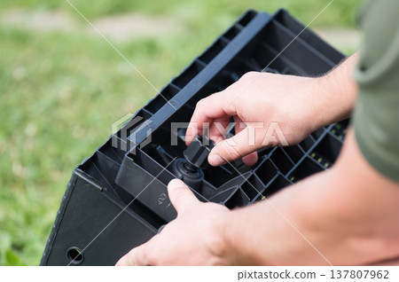 Assembling a black plastic crate outdoors. Close-up view of hands connecting components. Assembling a black plastic crate outdoors. Close-up view of hands connecting components. 137807962