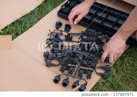 Assembling plastic parts outdoors. Man sorts black plastic components on cardboard. Assembling plastic parts outdoors. Man sorts black plastic components on cardboard. 137807971