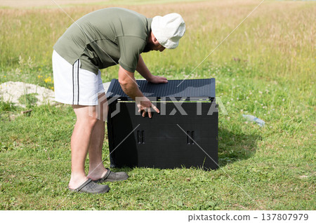Man assembling a black plastic storage unit outdoors on a grassy area. 137807979