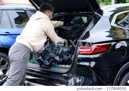Man loading black bags into open trunk of black SUV in parking lot on a rainy day in the afternoon Man loading black bags into open trunk of black SUV in parking lot on a rainy day in the afternoon 137808030
