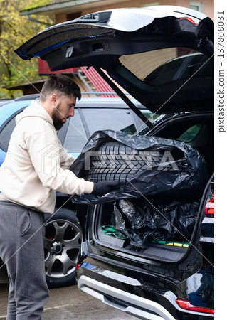 Man loading a tire into the trunk of a car at a location on a rainy day in an urban area 137808031