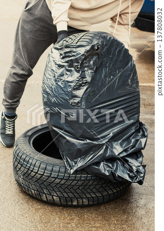 Person puts a black garbage bag on top of a tire in a parking area during daytime Person puts a black garbage bag on top of a tire in a parking area during daytime 137808032