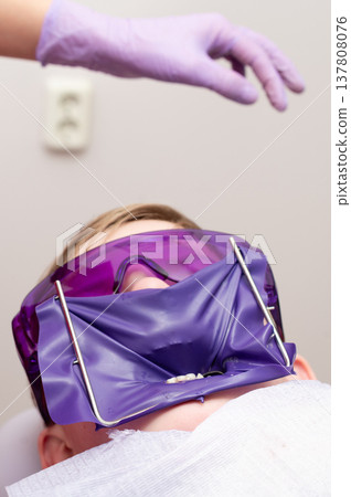 Young patient in a dental clinic prepares for treatment while wearing a protective face shield and goggles during an afternoon appointment in the care of a professional 137808076