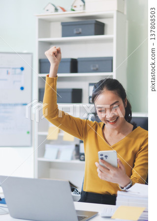 Asian woman works on laptop at office desk. She smiles happily while typing on keyboard near window with blinds. Professional woman works in business environment. Asian woman works on laptop at office desk. She smiles happily while typing on keyboard near window with blinds. Professional woman works in business environment. 137808503