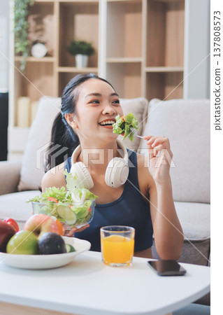 Fit young Asian women in activewear sitting on yoga mat and eating fresh vegetable salad after workout, representing clean eating, fitness lifestyle, wellness routine and health balance. 137808573