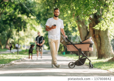 Smartphone in hand. Father is with cradle, having a walk in the park Smartphone in hand. Father is with cradle, having a walk in the park 137808642