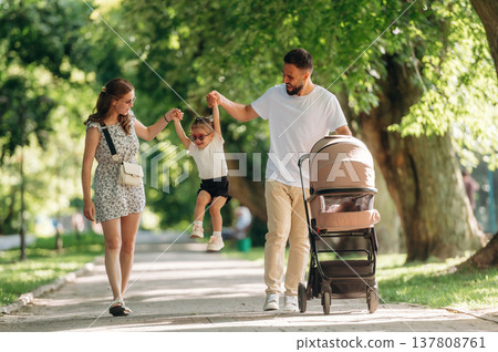 Holding girl by hands. Couple is with daughter and cradle are having a walk 137808761