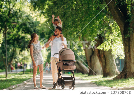 Girl is sitting on dad's shoulders. Couple is with daughter and cradle are having a walk Girl is sitting on dad's shoulders. Couple is with daughter and cradle are having a walk 137808768
