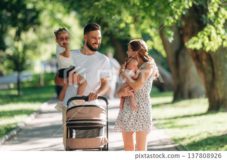 Baby is in hands. Couple is with daughter, newborn baby and cradle are having a walk Baby is in hands. Couple is with daughter, newborn baby and cradle are having a walk 137808926