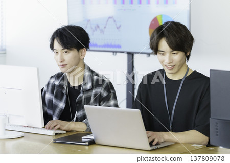 Two young Japanese men in casual clothes working on computers in an office. Two young Japanese men in casual clothes working on computers in an office. 137809078
