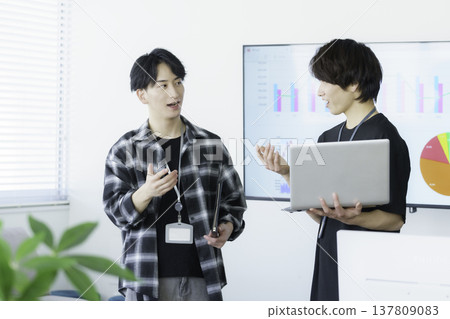 Two young Japanese men in casual clothes are talking, each holding a laptop. Two young Japanese men in casual clothes are talking, each holding a laptop. 137809083