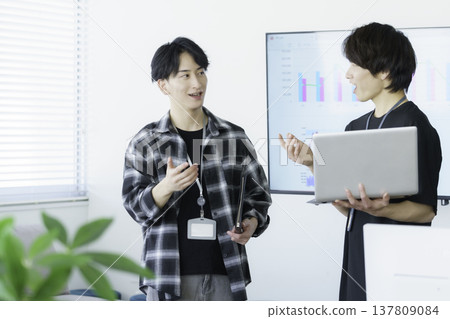 兩名身著便服的日本年輕男子正在交談，兩人各自拿著一台筆記型電腦。 137809084