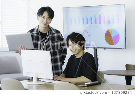 Two young Japanese men in casual clothes working on computers in an office. Two young Japanese men in casual clothes working on computers in an office. 137809284