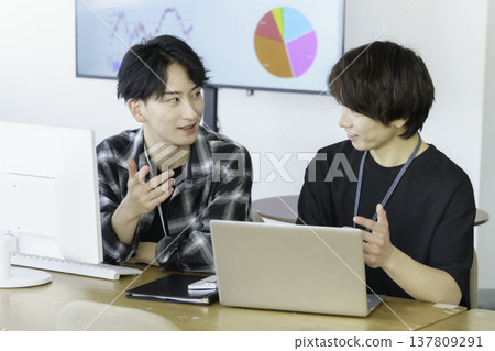 Two young Japanese men in casual clothes working on computers in an office. Two young Japanese men in casual clothes working on computers in an office. 137809291