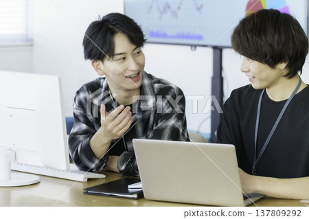 Two young Japanese men in casual clothes working on computers in an office. Two young Japanese men in casual clothes working on computers in an office. 137809292