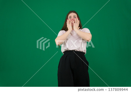 Various emotions of a European woman in a pink T-shirt and black pants on a green background with long brown hair in different poses 137809944