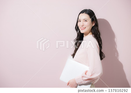 A cute Japanese woman in office casual attire, holding a laptop against a pink background, smiles and looks directly at the camera. 137810292