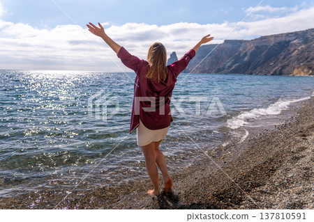 Woman beach freedom stands with arms raised facing the sparkling blue sea on a pebble shore near mountains Woman beach freedom stands with arms raised facing the sparkling blue sea on a pebble shore near mountains 137810591