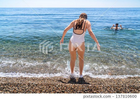Beach swimming woman walks into crystal clear sea water on a pebble beach during summer vacation under a bright sunny sky 137810600