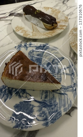 Slice of cheesecake on patterned plate with chocolate eclair in background Slice of cheesecake on patterned plate with chocolate eclair in background 137810676