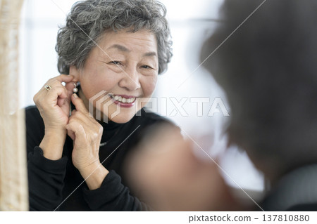 A smiling senior woman puts on earrings while looking in the mirror. 137810880